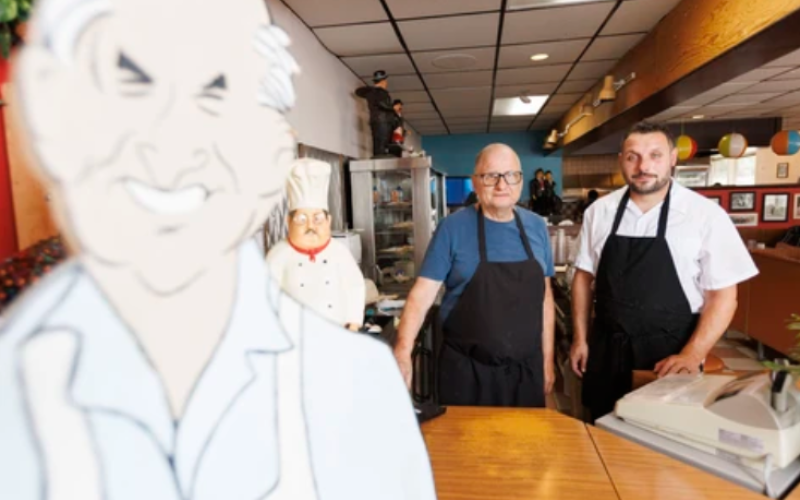 Leo Nikas, owner of Del-Mar Restaurant, and his son George pose with a caricature of Leo’s father at their London restaurant on Wednesday, Aug. 12, 2026. (Geoff Robins/The London Free Press)